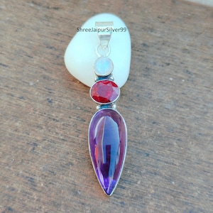 Puede incluir: Colgante de plata con tres piedras preciosas: una pequeña piedra redonda azul claro, una piedra roja ovalada y una gran piedra morada en forma de lágrima. El colgante tiene un lazo plateado en la parte superior. El texto "ShreejaipurSilver99" es visible.