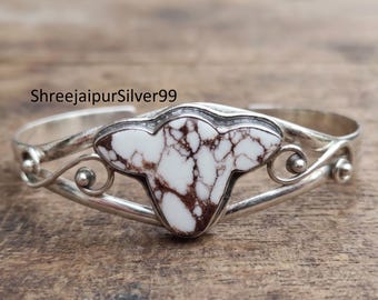 Peut inclure: Un bracelet manchette en argent orné d'une pierre blanche centrale en forme de bouclier avec des veines brunes. La bande métallique présente des volutes décoratives et de petites sphères en argent. Le texte ShreejaipurSilver99 est visible.
