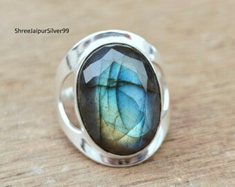 Peut inclure: Bague en argent ornée d'une pierre labradorite ovale et facettée. La pierre présente des reflets bleus, verts et jaunes. La bague a un design simple et moderne avec une finition polie. Le nom de la marque "ShreejaipurSilver99" est visible.