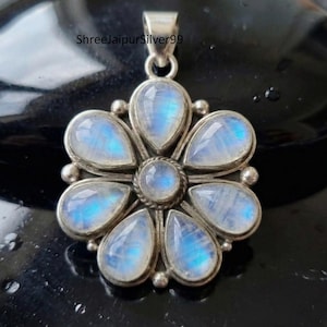 May include: A silver pendant featuring a flower design with seven teardrop-shaped moonstone gemstones. The moonstones have a blue and white iridescent glow. The pendant has a loop at the top for a chain. The text "ShreeJaipurSilver99" is visible.