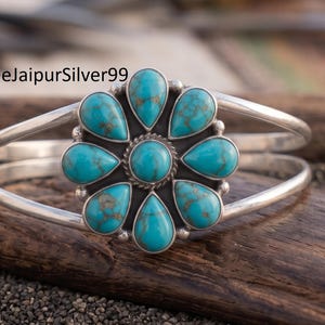 May include: A silver cuff bracelet featuring a central flower-shaped design composed of a round turquoise cabochon surrounded by eight teardrop-shaped turquoise stones. The metal has a polished finish, and the text ShreeJaipurSilver99 is visible.