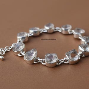 Puede incluir: Una pulsera de plata con piedras preciosas claras de forma irregular. La pulsera tiene un cierre de palanca. Las piedras están engastadas en biseles de plata y conectadas por pequeños eslabones de plata. El fondo es marrón.
