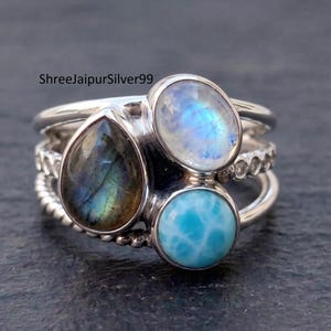 Pode incluir: Um conjunto de três anéis de prata empilháveis com pedras preciosas. Apresentam um labradorite em forma de gota, uma pedra da lua oval e um larimar redondo. As bandas dos anéis têm texturas diferentes, incluindo um design torcido.