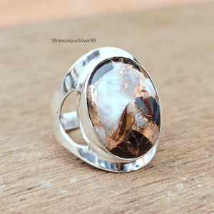 May include: A silver ring with an oval-shaped stone. The stone has a mix of colors, including white, brown, and black. The ring has a unique design with a cutout on each side. The text "ShreejaipurSilver99" is visible.