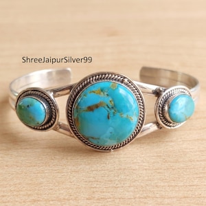 May include: A silver cuff bracelet with three turquoise stones. The central stone is the largest, encircled by a rope-style border. The bracelet has the text "ShreeJaipurSilver99".