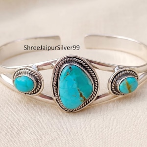 Könnte beinhalten: Ein silbernes Armreif-Armband mit drei Türkissteinen. Der zentrale Stein ist ein großes, unregelmäßiges Oval, flankiert von zwei kleineren, ovalen Türkissteinen. Das Armband hat ein geflochtenes Seildesign um den zentralen Stein und den Text "ShreeJaipurSilver99".