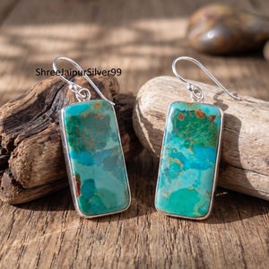 Puede incluir: Un par de pendientes colgantes rectangulares con piedras de turquesa pulidas con patrones de matriz marrón natural engastadas en marcos de plata. Los pendientes tienen ganchos de plata y el texto ShreeJaipurSilver99 es visible arriba.
