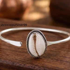 Puede incluir: Una pulsera de plata con un brazalete con una concha ovalada de color crema en el centro. La concha está enmarcada por un borde plateado retorcido. La pulsera descansa sobre una superficie de madera oscura.