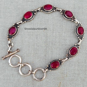 Puede incluir: Un brazalete de plata con cierre de palanca y piedras preciosas rojas de forma ovalada. El brazalete está hecho de plata y tiene un acabado pulido.