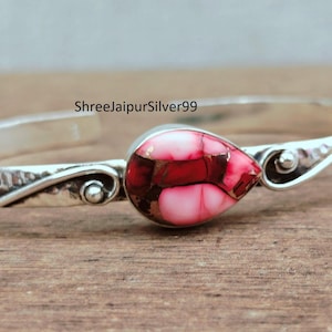 May include: A silver bangle bracelet with a teardrop-shaped centerpiece. The centerpiece features a mosaic of red and pink stones set in a silver bezel. The bracelet has a hammered texture.