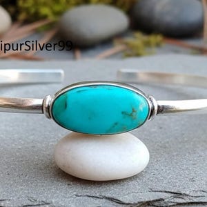 May include: A silver cuff bracelet featuring a central oval turquoise stone. The turquoise is set in a silver bezel, and the bracelet has a simple, elegant design. The bracelet is displayed on a white stone.