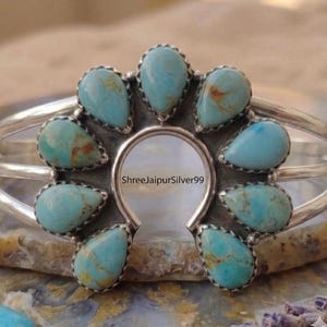 May include: A silver cuff bracelet featuring a central horseshoe-shaped frame adorned with eight teardrop-shaped turquoise stones. The stones display light blue hues with brown matrix veining. The text ShreeJaipurSilver99 is visible in the center.