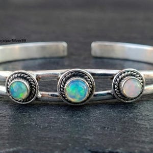 May include: A silver cuff bracelet featuring three opal gemstones. Each opal is set in a circular frame with a twisted rope design. The bracelet has a double-band design and is open at the ends.