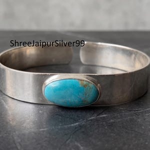 Könnte beinhalten: Ein silbernes Armband mit einer glatten, polierten Schiene und einem zentralen, ovalen Türkis-Edelstein mit natürlichen braunen Maserungen. Der Text ShreeJaipurSilver99 ist oben links über dem Metallband im Bild zu sehen.