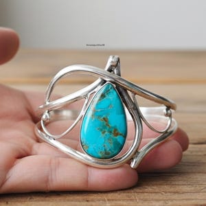 May include: A silver cuff bracelet with a large, teardrop-shaped turquoise stone. The stone is a vibrant blue-green colour with brown matrix patterns. The bracelet has an open design with curved, flowing silver elements.