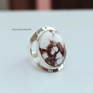 May include: A silver ring featuring an oval-shaped white stone with brown marbling. The ring has a wide band with a cutout design on each side of the stone. The text "ShreejaipurSilver99" is visible in the image.