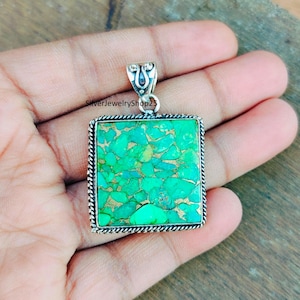 May include: A square pendant with a green turquoise stone set in silver. The stone has a mottled pattern of green and gold.