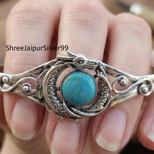 May include: A silver cuff bracelet featuring a central round turquoise gemstone encircled by a detailed dragon motif with scales and a head. The metal band displays polished silver surfaces with decorative scrollwork and swirling accents.
