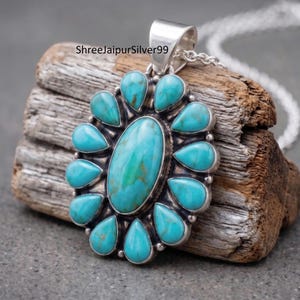 May include: A sterling silver pendant featuring a large central oval turquoise stone surrounded by twelve smaller teardrop-shaped turquoise stones. The metal setting has a polished finish with a bail at the top and a silver chain attached.