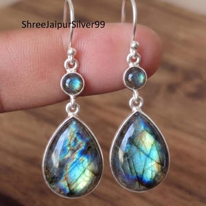 Puede incluir: Un par de pendientes de plata con una pequeña piedra de labradorita redonda sobre una piedra de labradorita en forma de pera. Las piedras muestran destellos azules y verdes iridiscentes en los engastes de plata. El texto ShreeJaipurSilver99 es visible.