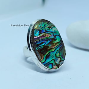 May include: A silver ring with an oval-shaped abalone shell inlay. The shell has a blue, green, and purple iridescent pattern.