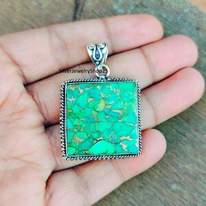 May include: A square pendant with a green turquoise stone set in silver. The stone has a mottled pattern of green and gold.