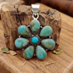 May include: A silver pendant shaped like a flower with six teardrop-shaped turquoise stones surrounding a small, round, bright blue turquoise center stone. The metal setting is polished silver, and the text ShreeJaipurSilver99 is visible.