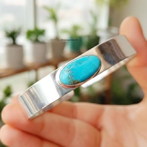 May include: A silver cuff bracelet with a bright turquoise stone. The oval stone is set in a bezel setting, and the bracelet has a polished, rectangular design. The turquoise stone has a blue-green hue with brown matrix.