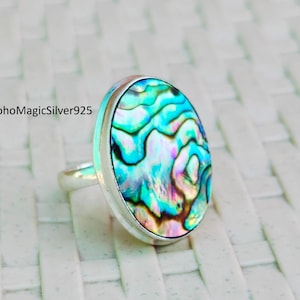 Puede incluir: Un anillo de plata con una incrustación redonda de concha de abulón. La concha tiene un patrón azul, verde e iridiscente.