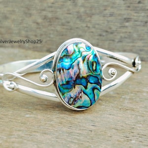 Puede incluir: Un brazalete de plata con una gran incrustación ovalada de concha de abulón. La concha tiene colores iridiscentes azules, verdes y morados.