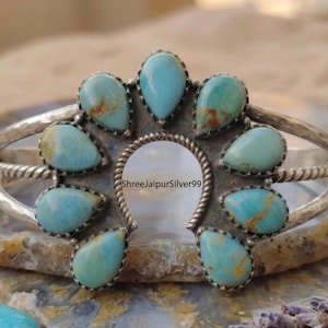 May include: A silver cuff bracelet featuring a central horseshoe-shaped frame adorned with nine teardrop-shaped turquoise stones. The metal band has a hammered texture and twisted wire detailing. The text ShreeJaipurSilver99 is visible.