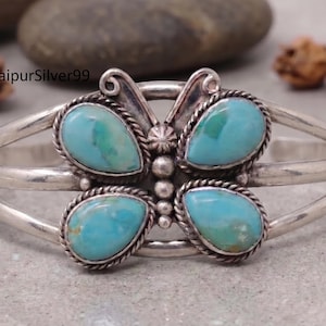 May include: A silver cuff bracelet featuring a butterfly-shaped centerpiece with four teardrop-shaped turquoise stones. The stones are set in twisted wire borders, and the center of the butterfly is accented with small silver beads and metal coils.