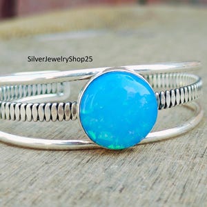 May include: A silver bangle bracelet featuring a blue opal gemstone. The bracelet has a textured design and is made of two bands.