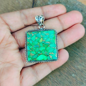 May include: A silver pendant with a square turquoise stone. The stone is a bright green color and has a mottled pattern. The pendant is on a hand.