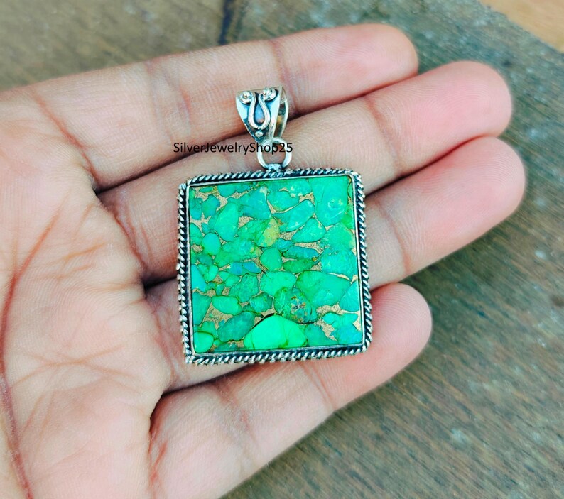 May include: A silver pendant with a square turquoise stone. The stone is a bright green color and has a mottled pattern. The pendant is on a hand.