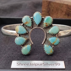 Pode incluir: Uma pulseira de prata com um design central em forma de ferradura adornado com nove pedras de turquesa em forma de gota com matriz castanha. O centro da ferradura tem um detalhe de fio de prata torcido. Etiqueta metálica: ShreeJaipurSilver99.