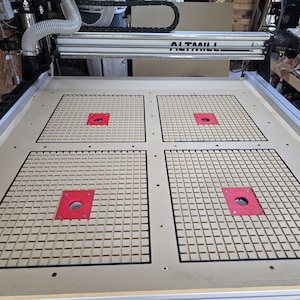 以下が含まれることがあります： 明るい木材の作業面を持つCNCマシン。表面には、格子模様と赤いインサートが付いた4つの正方形のセクションがあります。マシンのフレームはシルバーで、「ALTAMILL」という名前が見えます。