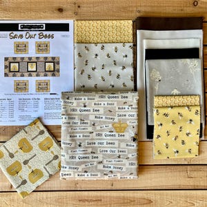 May include: A collection of bee-themed fabric pieces, including patterned fabrics with bees, honeycombs, and text like "Save Our Bees" and "Make Honey." There's also a pattern sheet with bee-related artwork, all on a wooden surface.