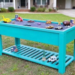 Peut inclure: Une table de jeu en bois turquoise vif avec un plateau rectangulaire doté d'une surface bleu foncé et de marquages routiers blancs. La table comprend une étagère inférieure à lattes et est décorée de petites voitures, de blocs et d'un ballon.
