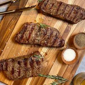May include: Three grilled steaks with grill marks, garnished with rosemary, rest on a wooden cutting board. Small wooden bowls hold salt and pepper. Gold tongs are visible in the upper left corner. The steaks are a rich brown color.