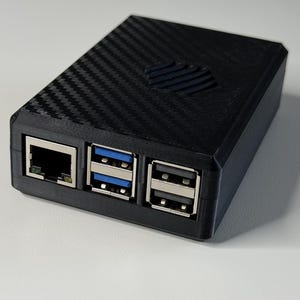 May include: A black rectangular electronic device case with a textured carbon fiber pattern. The case has an Ethernet port and two USB ports. The top of the case has a small, circular design. The device is on a white surface.