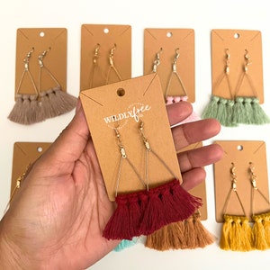 May include: A set of triangle-shaped earrings with gold-tone metal hoops and colorful tassel fringe. The earrings are displayed on brown card stock with the text "WILDLYfree" printed on the card.