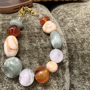 May include: A phone charm featuring a series of smooth, rounded beads in shades of sage green, amber, cream, and white. The beads are strung together and attached to gold-toned metal lobster clasps, creating a decorative accessory for a mobile device.