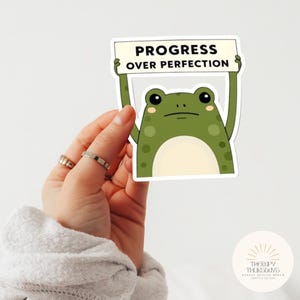 May include: A sticker featuring a cartoon frog holding a sign that reads "PROGRESS OVER PERFECTION." The frog is green with darker green spots and a neutral expression. The sticker is held by a hand with rings.