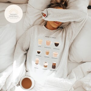 Puede incluir: Una sudadera gris con el texto "ANXIETY JUICE" e ilustraciones de diferentes bebidas de café, incluyendo latte, cappuccino, americano, espresso, cortado, macchiato, mocha y café helado.