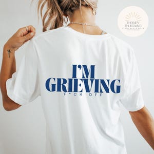 May include: White t-shirt with blue text that reads "I'm Grieving F*ck Off"