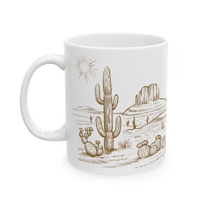 Peut inclure: Une tasse en céramique blanche avec un dessin au trait brun d'un paysage désertique avec un grand cactus, des cactus plus petits et une chaîne de montagnes. Le soleil brille dans le ciel.