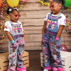 May include: Two young girls wearing matching denim overalls with colorful doll graphics and pink fur trim. The overalls have the text "Messiah" and "Sovanna" on them. They are wearing white t-shirts and sunglasses, posing in front of a wooden backdrop with balloons.