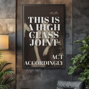 May include: A framed black and white print with the text "THIS IS A HIGH CLASS JOINT ACT ACCORDINGLY." The print hangs on a dark gray textured wall. A modern chair and side table are in the foreground.