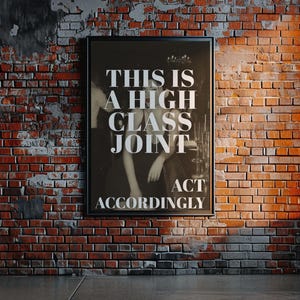 May include: A framed black and white poster with the text "THIS IS A HIGH CLASS JOINT ACT ACCORDINGLY" hangs on a brick wall. The brick wall is red and brown, with a concrete floor. A window is visible on the right.